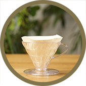 A circular image of a clear Hario V60 with a filter paper inside of it. The V60 is sitting on a wooden table.